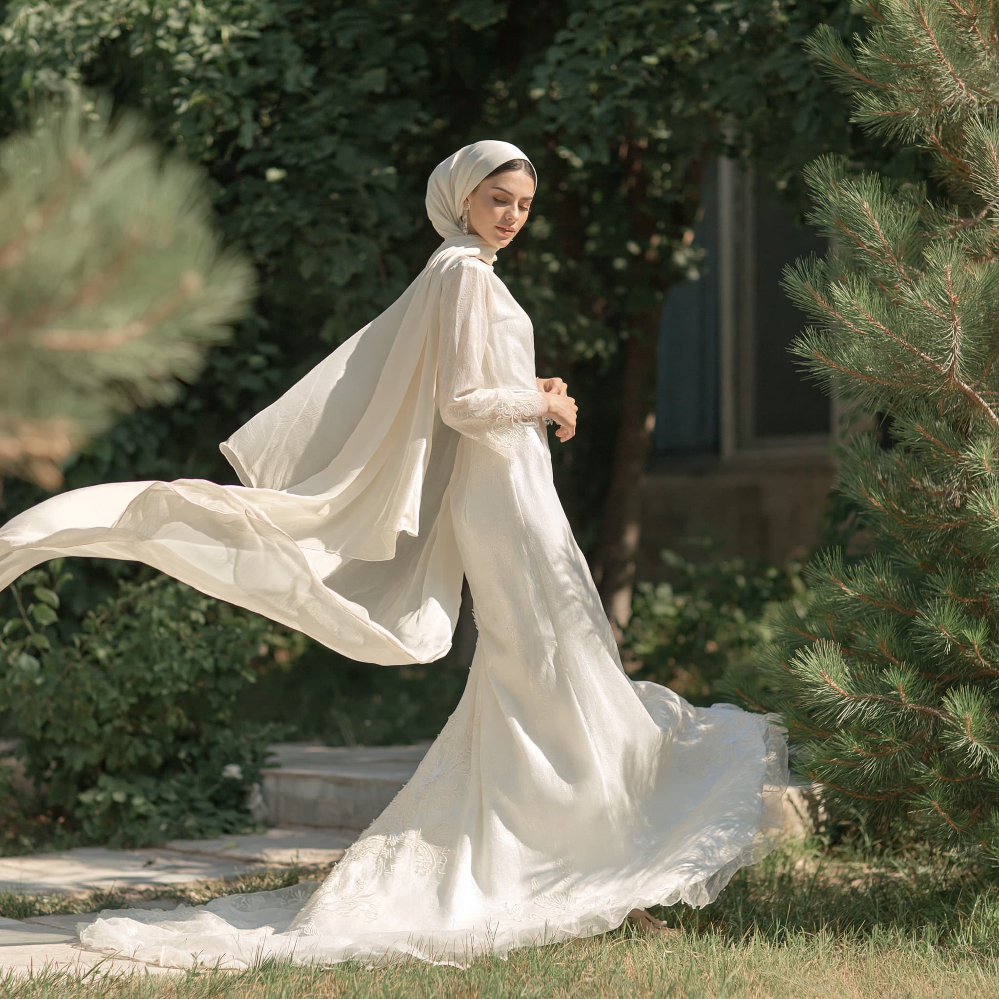 A woman in a flowing white Safid gown and scarf, walking through a sunlit garden